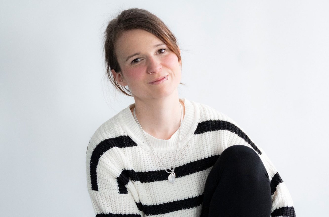 Laura Bartels.jpg Junge Frau mit lässigem Streifensweater sitzt lächelnd vor hellem Hintergrund.